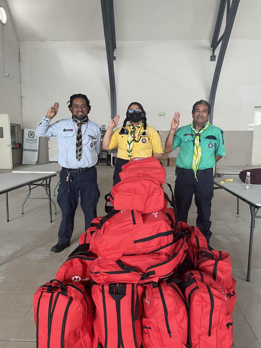 Tres integrantes de la Asociación de Scouts de México junto a un grupo de mochilas rojas